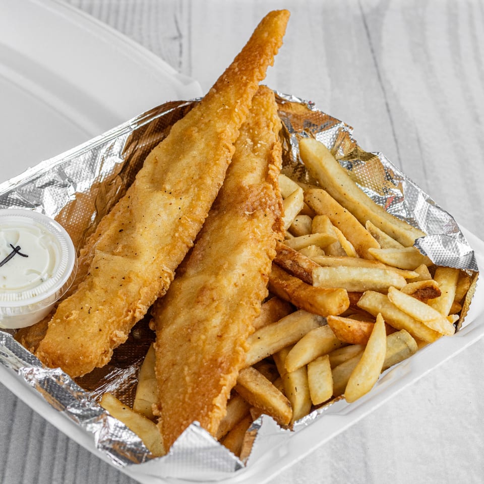 Two pieces of fried fish and french fries in a foil-lined takeout container with a side of tartar sauce.