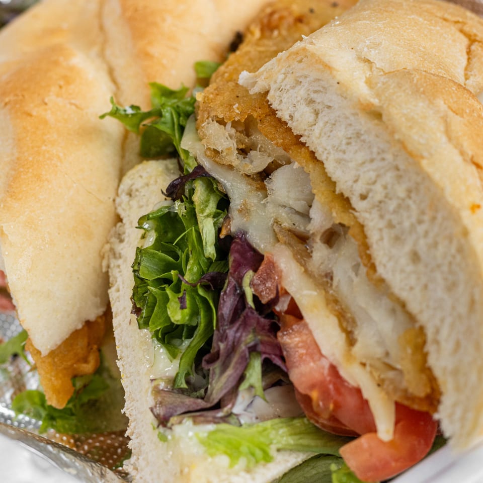 Close-up of a Corbos fried fish sandwich with lettuce, tomatoes, and melted cheese on a bun.
